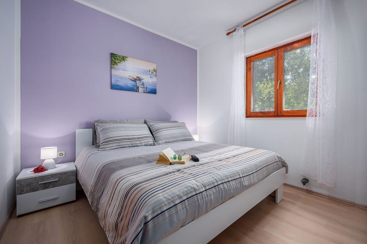 A bedroom with a bed, a nightstand, a lamp, a window, and a painting on the wall.
