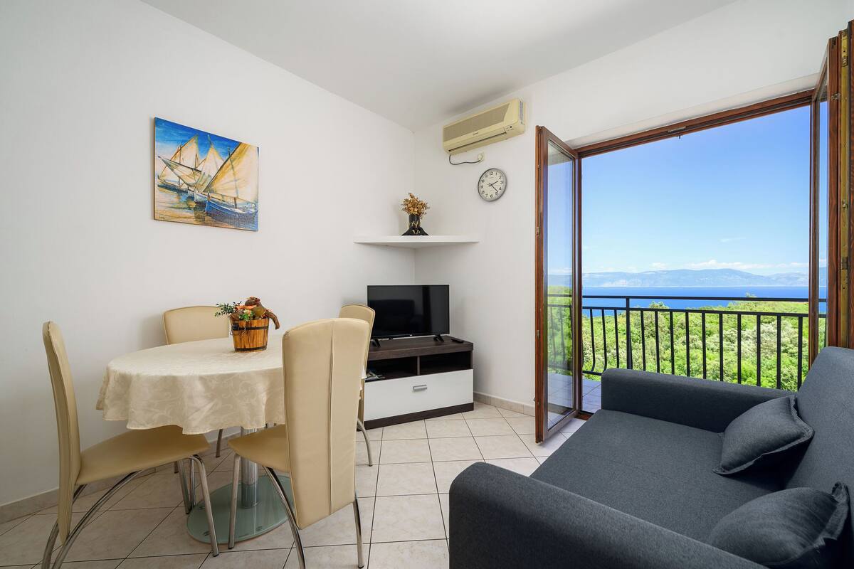 A balcony with a view of mountains, a dining area with a table and chairs, a sofa, a TV, and a painting on the wall.
