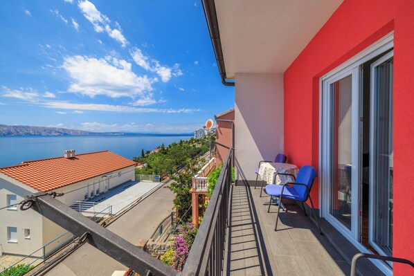 A balcony with a view of the sea, a red building, and a blue chair.