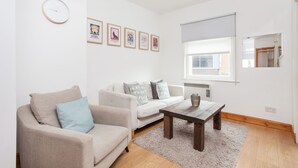 A modern living room with a beige sofa, a wooden coffee table, and a large window with blinds. - Basic Apartment for Two Amazing Location (London)