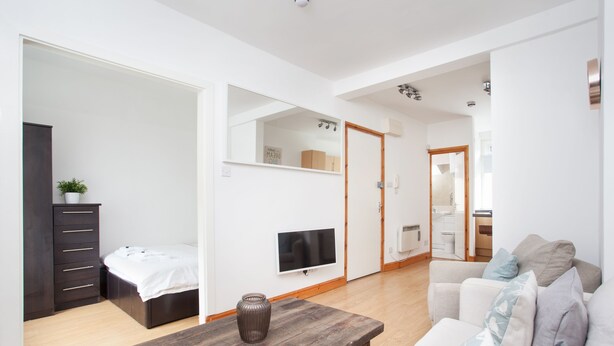 A modern living room with a wooden coffee table, a white sofa with patterned cushions, a TV mounted on the wall, and a small plant on a dark wooden dresser. - Basic Apartment for Two Amazing Location (London)