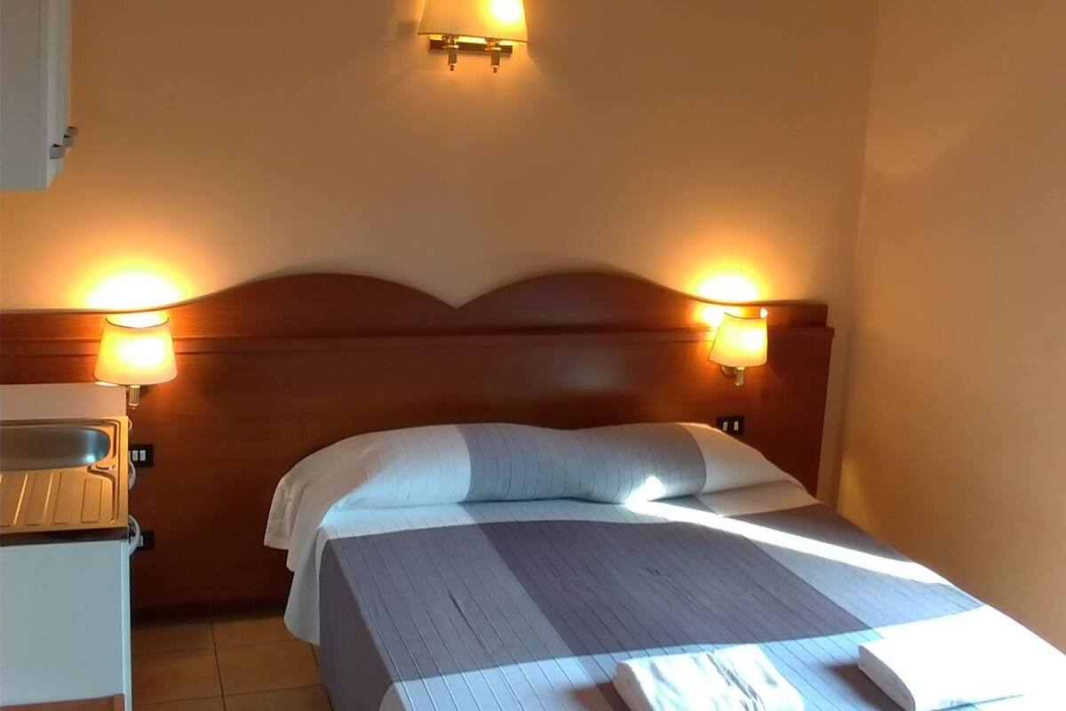 Quarto de hotel com uma cama, duas luminárias laterais, um ventilador de teto e uma mesinha.