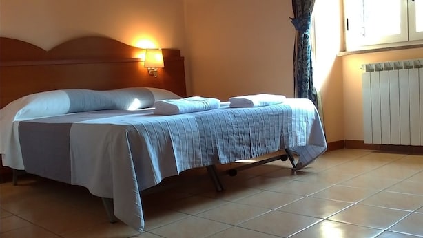A hotel room with a bed, a headboard, a wall-mounted lamp, a window with curtains, and a radiator. - Near Termini Station (Rome)