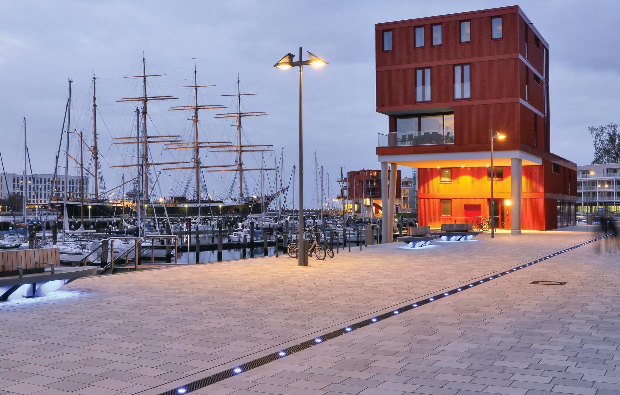 Un edifici modern amb façana vermella, una zona exterior coberta amb seients i una marina amb velers al fons.
