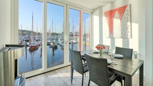 A dining area with a view of a marina through large windows, wicker chairs, a table set with plates and glasses, and a red and white abstract painting on the wall. - Passatvilla - Apartment 2 (Travemünde Waterfront)