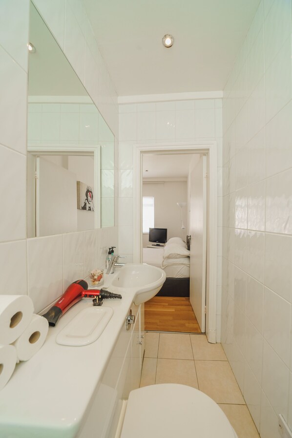 Un baño moderno con un lavamanos blanco, un espejo y un inodoro. Hay un secador de pelo rojo y una botella de champú sobre el lavamanos.