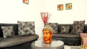 Un salon avec un canapé noir, des coussins à motifs, une table basse ronde en verre et une statue de Bouddha.