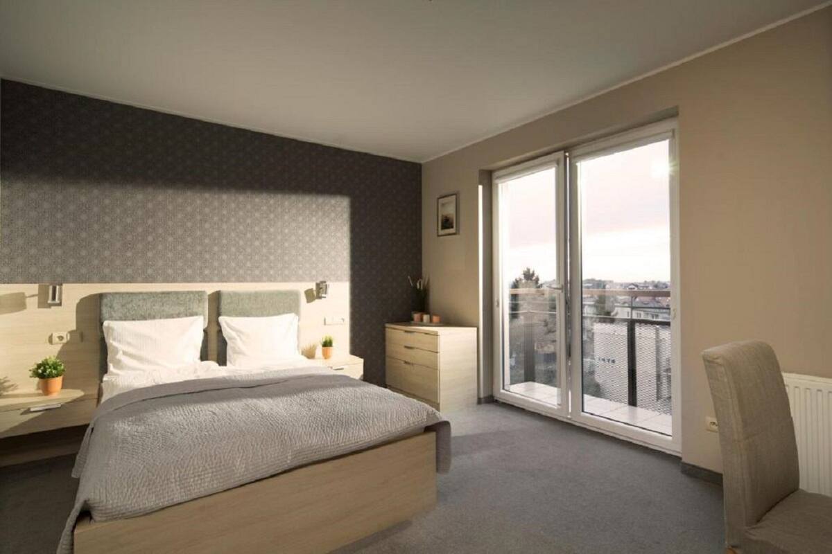 A modern bedroom with a large bed, a nightstand, a dresser, and a chair. There is a large window providing a view of the city.