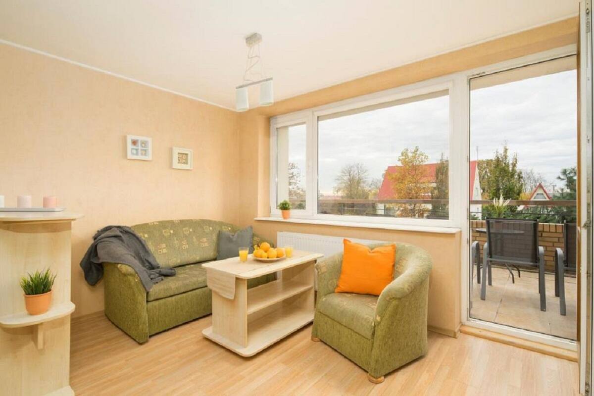 A cozy living room with a green sofa, a small wooden table with fruit, and a window overlooking a patio.