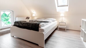 A modern bedroom with a sloped ceiling, a bed with a dark throw, two bedside lamps, and a window with a view of greenery. - Beautiful apartment "chicken coop" in the countryside (Harrierwurp)