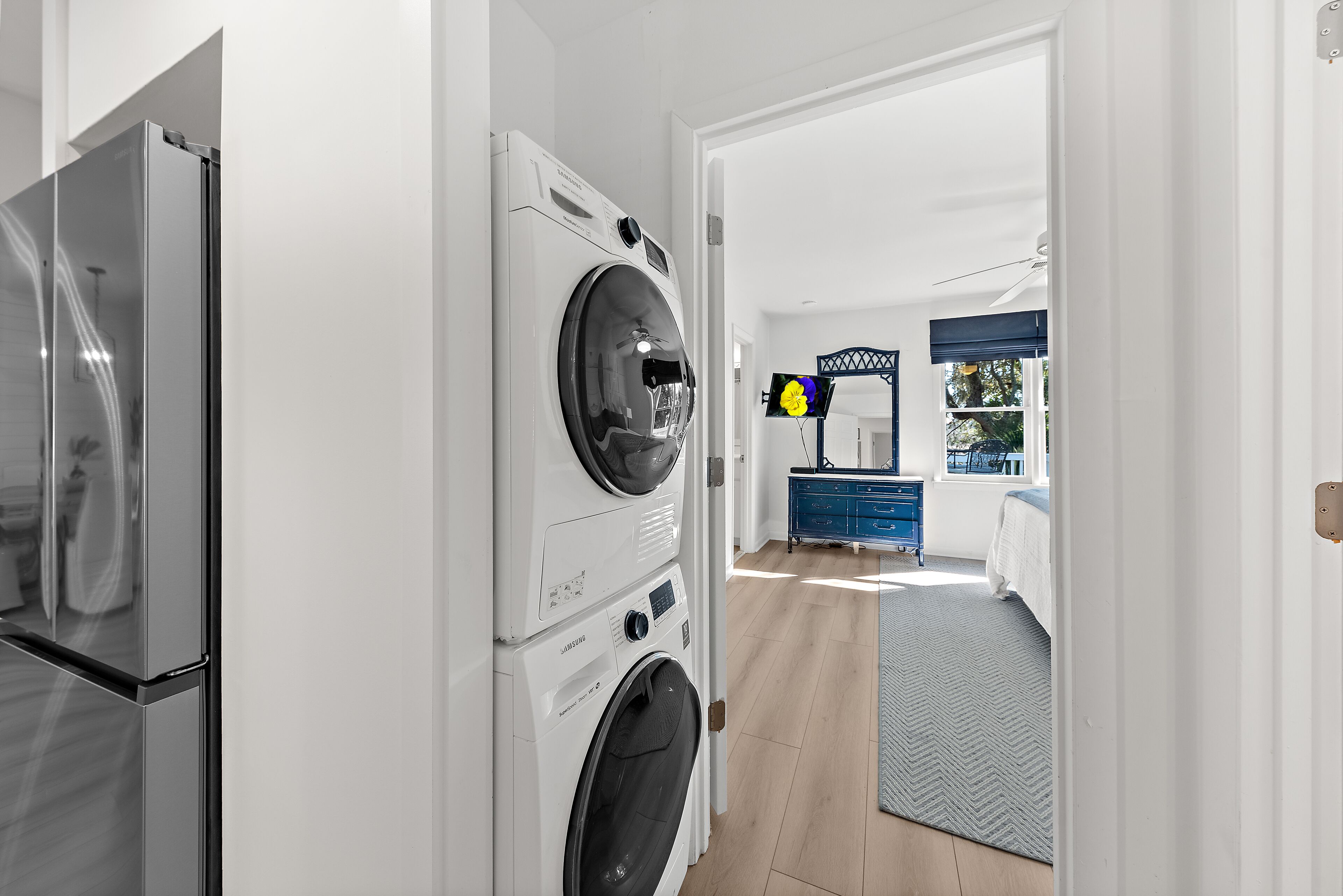 A laundry room with a front-loading washer and dryer, a blue dresser, and a bed with a patterned bedspread in the adjacent bedroom.