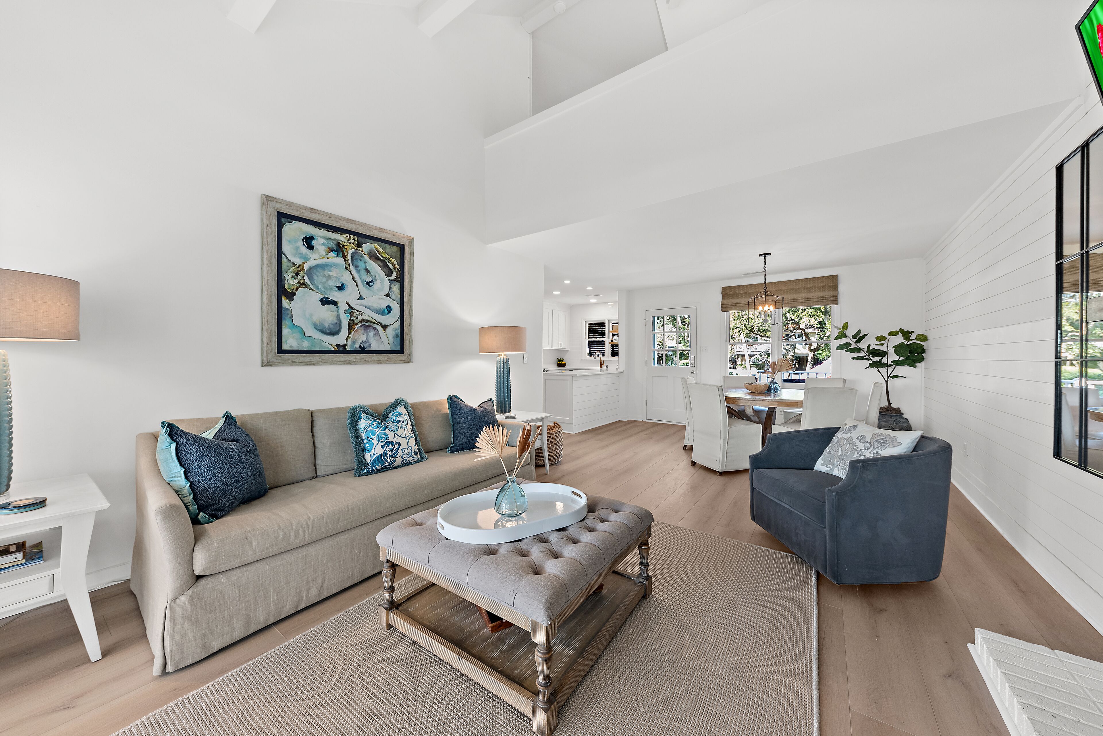 Ein geräumiges Wohnzimmer mit einem beigefarbenen Sofa, einem grauen Hocker und einem blauen Sessel. An der Wand hängt ein Gemälde und im Hintergrund ist ein Essbereich zu sehen.