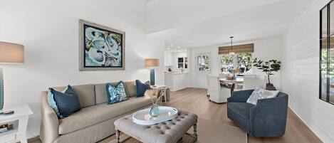Ein geräumiges Wohnzimmer mit einem beigefarbenen Sofa, einem grauen Hocker und einem blauen Sessel. An der Wand hängt ein Gemälde und im Hintergrund ist ein Essbereich zu sehen.