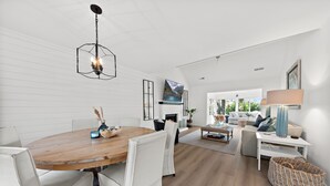 A spacious living area with a wooden dining table, white upholstered chairs, a fireplace, and a large window offering a view of the outdoors. - Pet Friendly 3BR Tarpon Pond Cottage! Close to Beach/Pools (Seabrook Island)