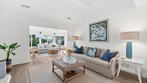 A modern living room with a sofa, ottoman, and side table, a large abstract painting, and a potted plant. - Pet Friendly 3BR Tarpon Pond Cottage! Close to Beach/Pools (Seabrook Island)
