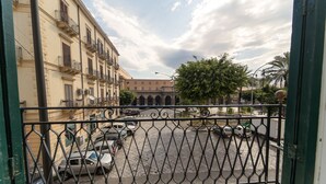 Blick von einem Balkon mit schmiedeeisernem Geländer auf eine Straße. Es sind Gebäude mit Balkonen, parkende Autos und ein klarer Himmel zu sehen.