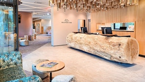 Un espace d’accueil moderne comprenant un comptoir de réception en bois, un grand banc en forme de tronc d’arbre et un coin salon avec des chaises à motifs.
