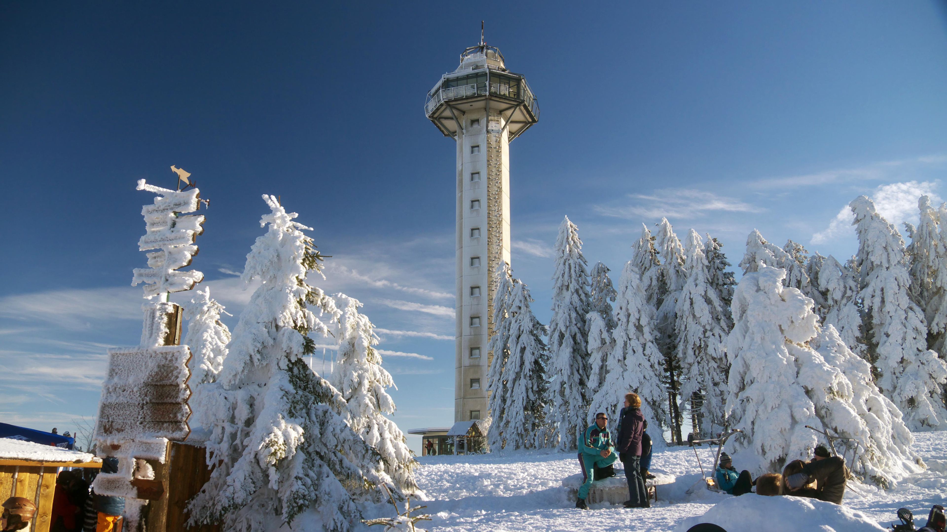 A snow-covered landscape with a tall tower structure, surrounded by snow-laden trees and a clear sky.