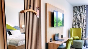 Une chambre d’hôtel avec un grand miroir, une cloison en lattes de bois, un bureau avec un ordinateur portable, une chaise verte et un lit avec un oreiller vert.
