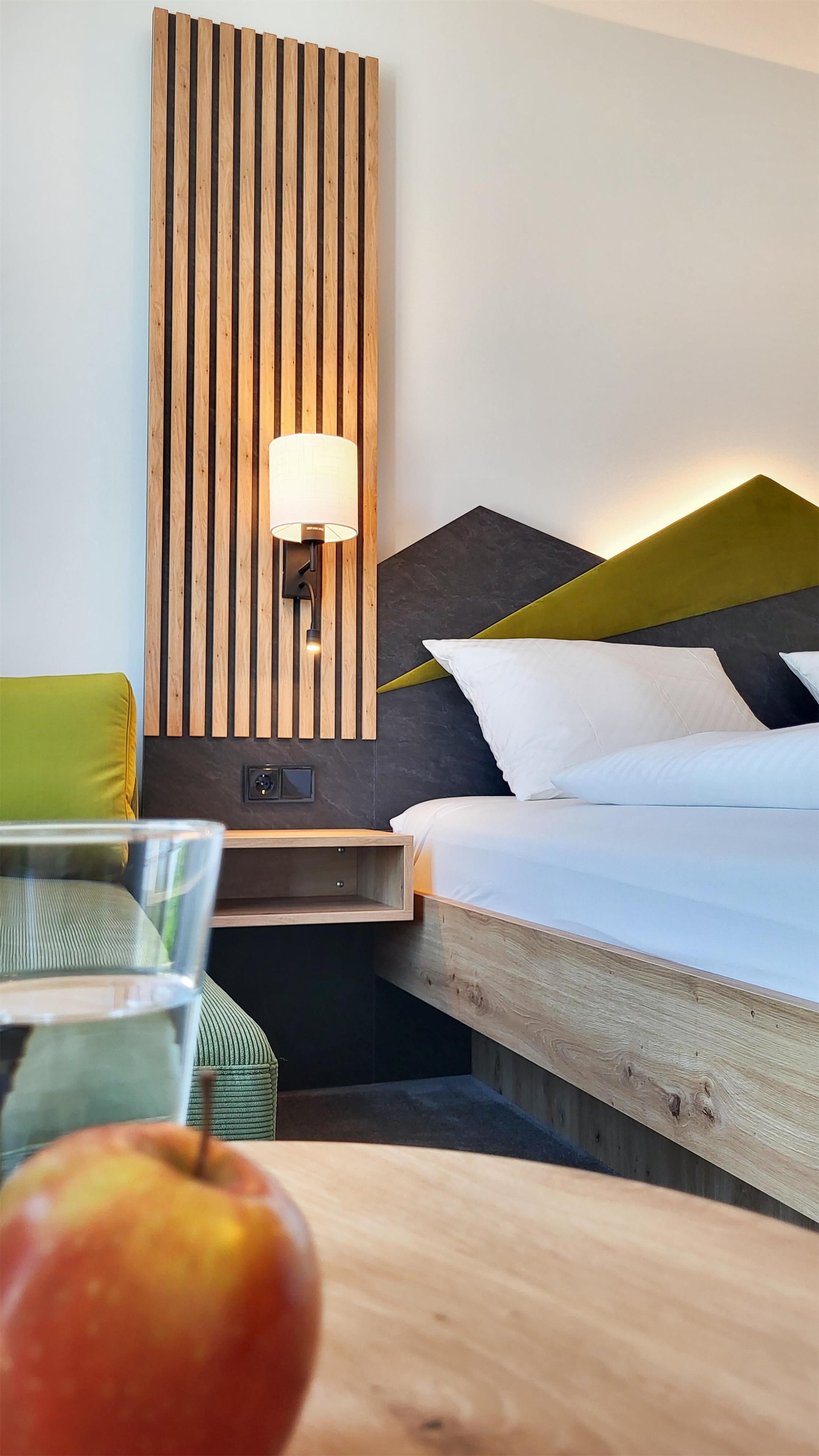 A hotel room with a wooden slat wall, a bedside table with a lamp, a bed with pillows, and a green chair.