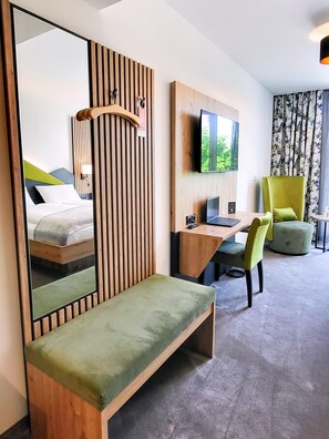 Ein Hotelzimmer mit Bett, Schreibtisch, Stuhl, einem großen Spiegel, einem Fernseher und einem Fenster mit Vorhängen.