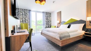 Ein modernes Hotelzimmer mit einem hölzernen Bett, einem Schreibtisch mit einem Laptop, einem grünen Sessel und einem großen Fenster mit Vorhängen.