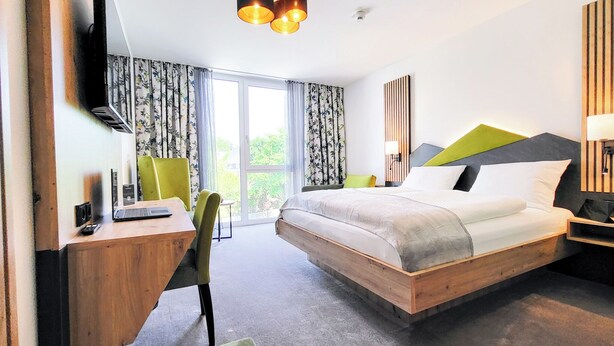 Una habitación de hotel moderna con una cama de madera, un escritorio con una computadora portátil, una silla verde y un ventanal grande con cortinas.