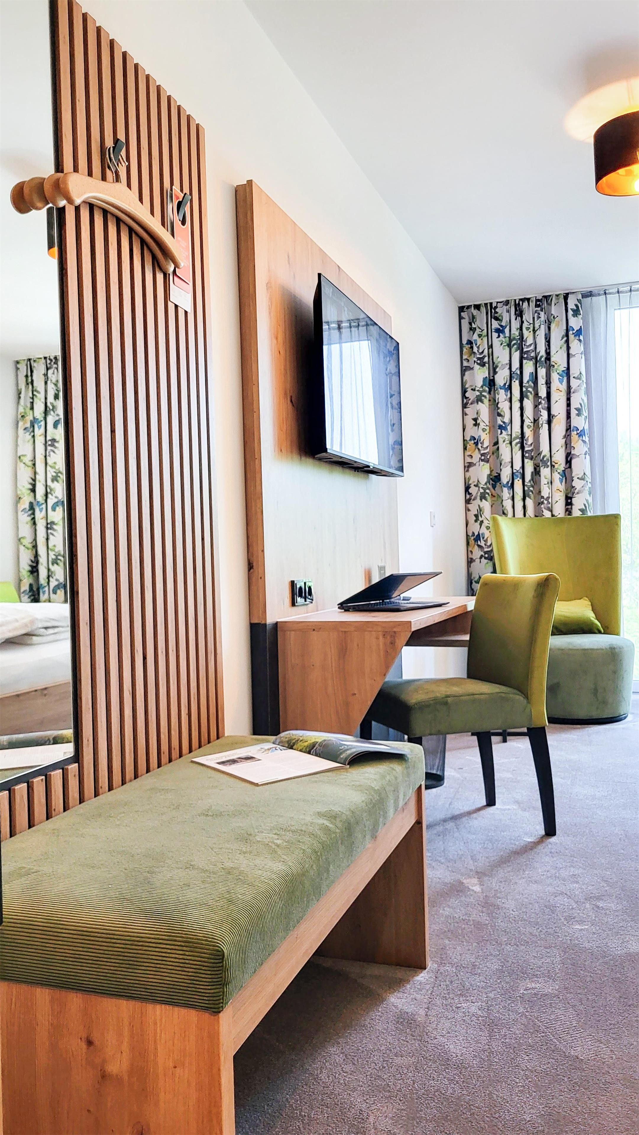 A modern hotel room with a wooden bench, a green sofa, a flat-screen TV mounted on the wall, and a window with floral curtains.