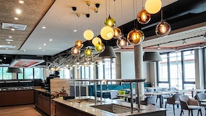 Un intérieur de restaurant moderne avec un îlot central, des luminaires suspendus et une vue sur l’extérieur grâce à de grandes fenêtres.