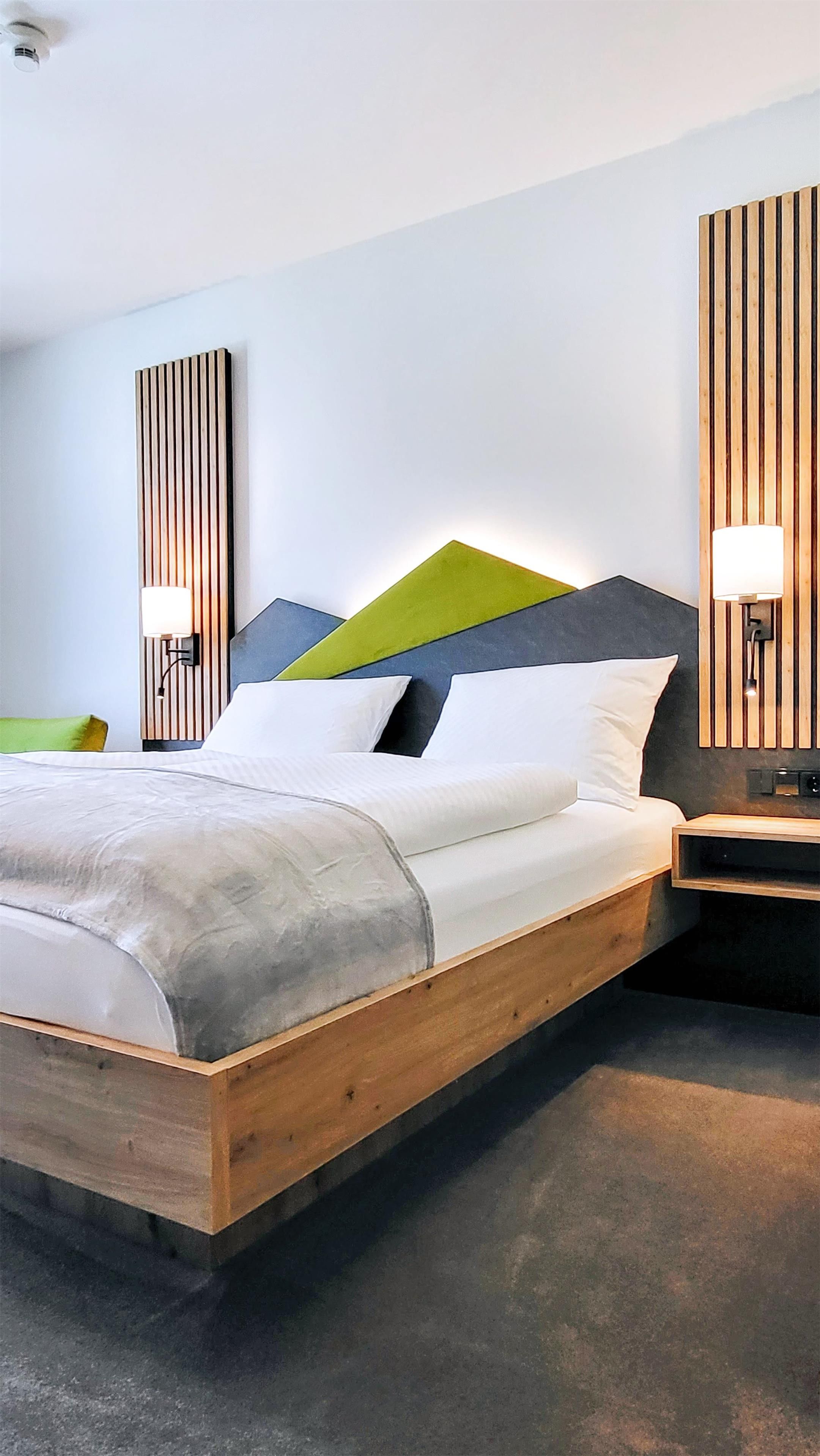 A modern hotel room with a wooden bed frame, a geometric-patterned headboard, and a bedside table with a lamp.