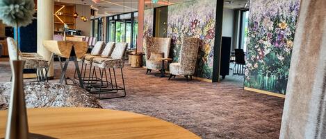 A hotel lobby with a carpeted floor, seating area with chairs, a wooden table, and a large floral mural on the wall.