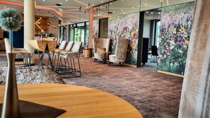 Un hall de hotel con piso alfombrado, zona de estar con sillas, una mesa de madera y un gran mural floral en la pared.