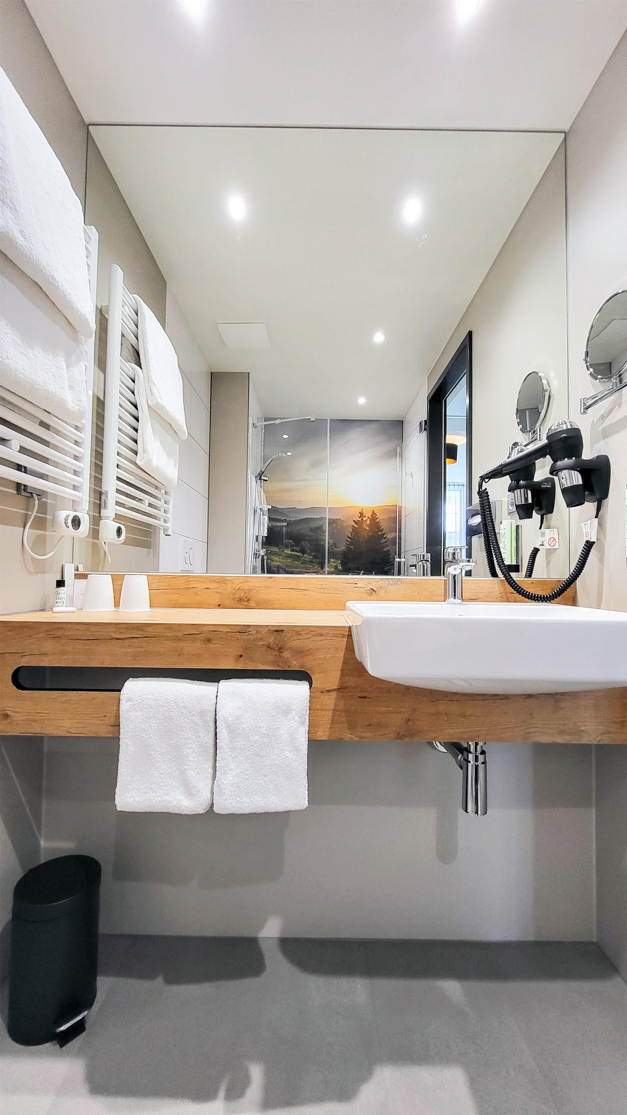 A modern bathroom with a wooden vanity, a large mirror, and a view of a sunset through the glass wall.