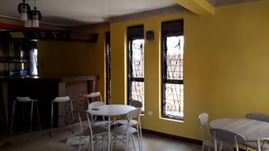 A dining area with a white table and chairs, a bar area with stools, and two windows with decorative iron bars. - Egret Pilgrims Inn (Kampala)