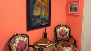 A room with a vibrant orange wall, a framed painting of flowers, two ornate chairs, and a small table with a book.