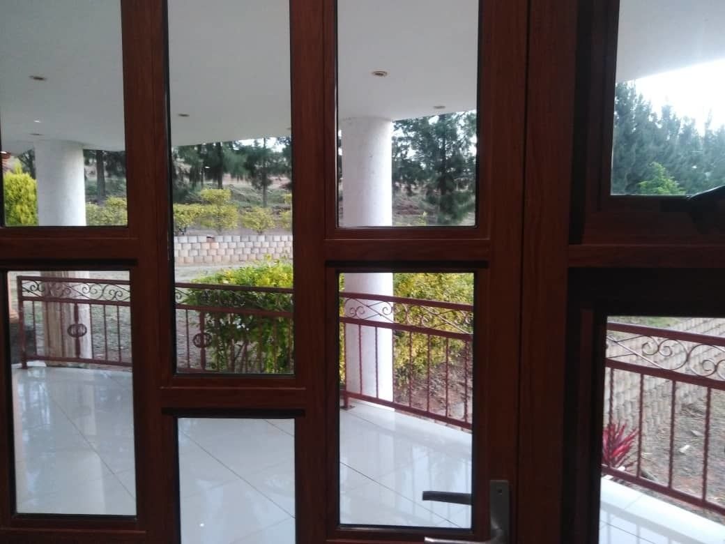 A view through a wooden-framed window with a metal railing, leading to an outdoor area with a tiled floor and greenery.