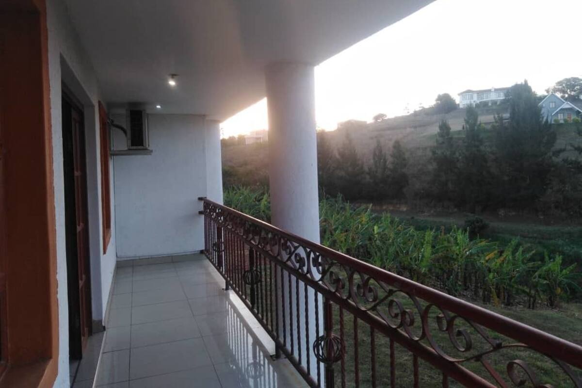 A balcony with a metal railing, white pillars, and a view of a green landscape with trees and a building in the distance.