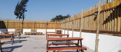 Un patio al aire libre con bancos y mesas de madera, rodeado por una valla de madera y una pared blanca.