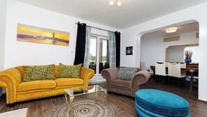 Un soggiorno con un divano giallo, una poltrona marrone, un tavolino in vetro e un poggiapiedi blu. Si nota un quadro raffigurante un faro appeso al muro, un'area pranzo con uno specchio e un tappeto decorato sul pavimento.