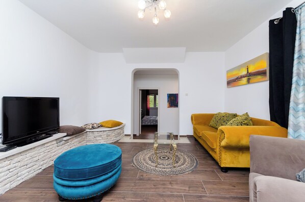 A living room with a flat-screen TV, a yellow sofa, a blue ottoman, and a patterned rug.