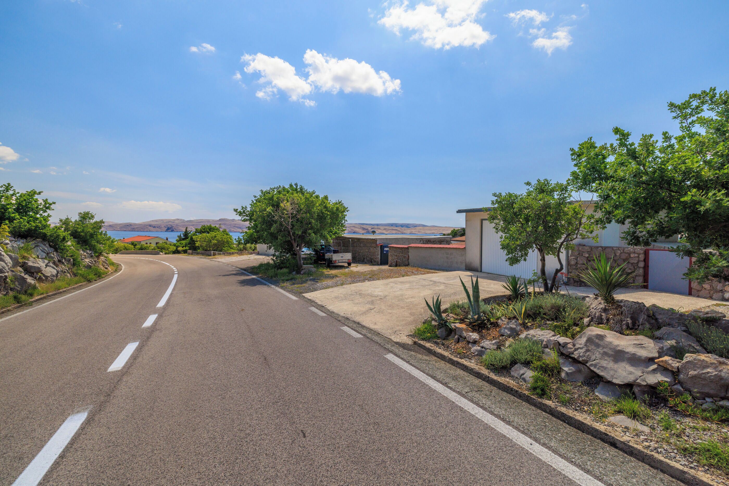 A curved road with a clear blue sky, a small building with a red roof, and a rocky garden area.