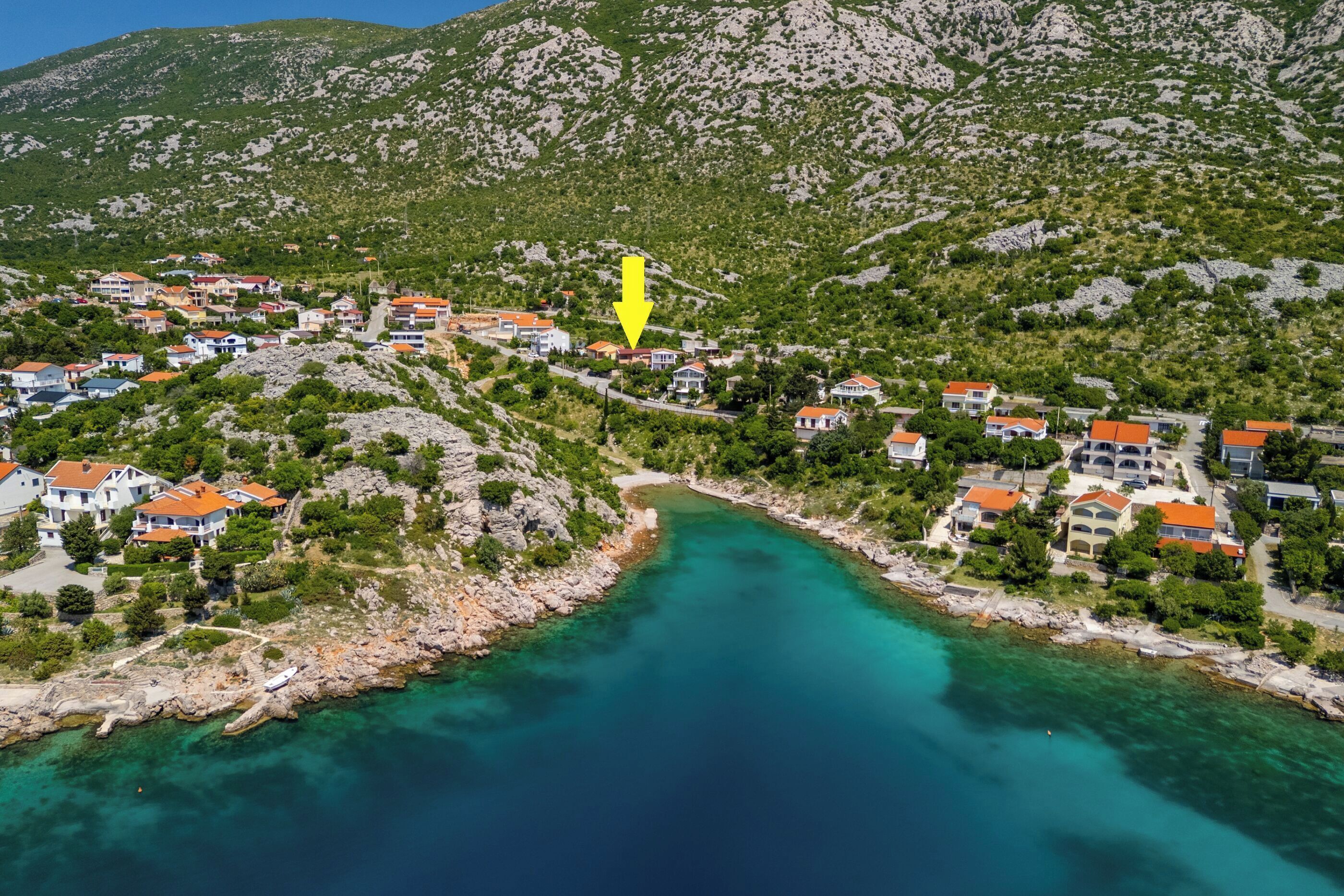 A coastal village with houses along the shoreline, a clear turquoise sea, and a mountainous backdrop.