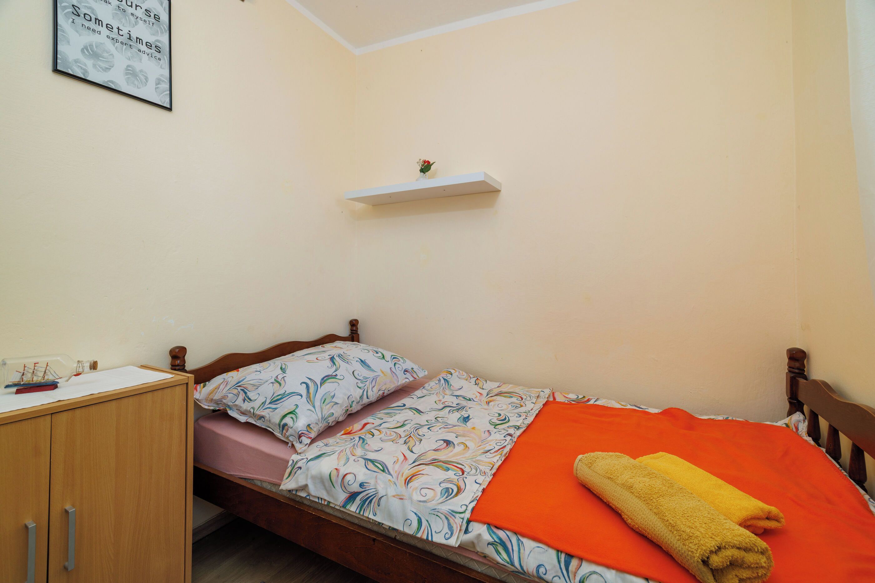 A small, neatly arranged bedroom with a single bed, a wooden nightstand, a small shelf, and a framed quote on the wall.