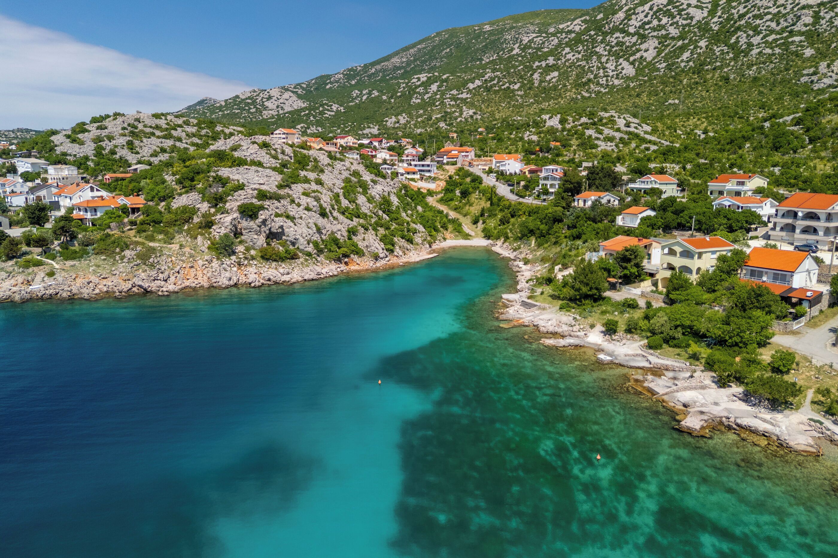A coastal village with houses on a hillside, a clear turquoise sea, and rocky terrain.