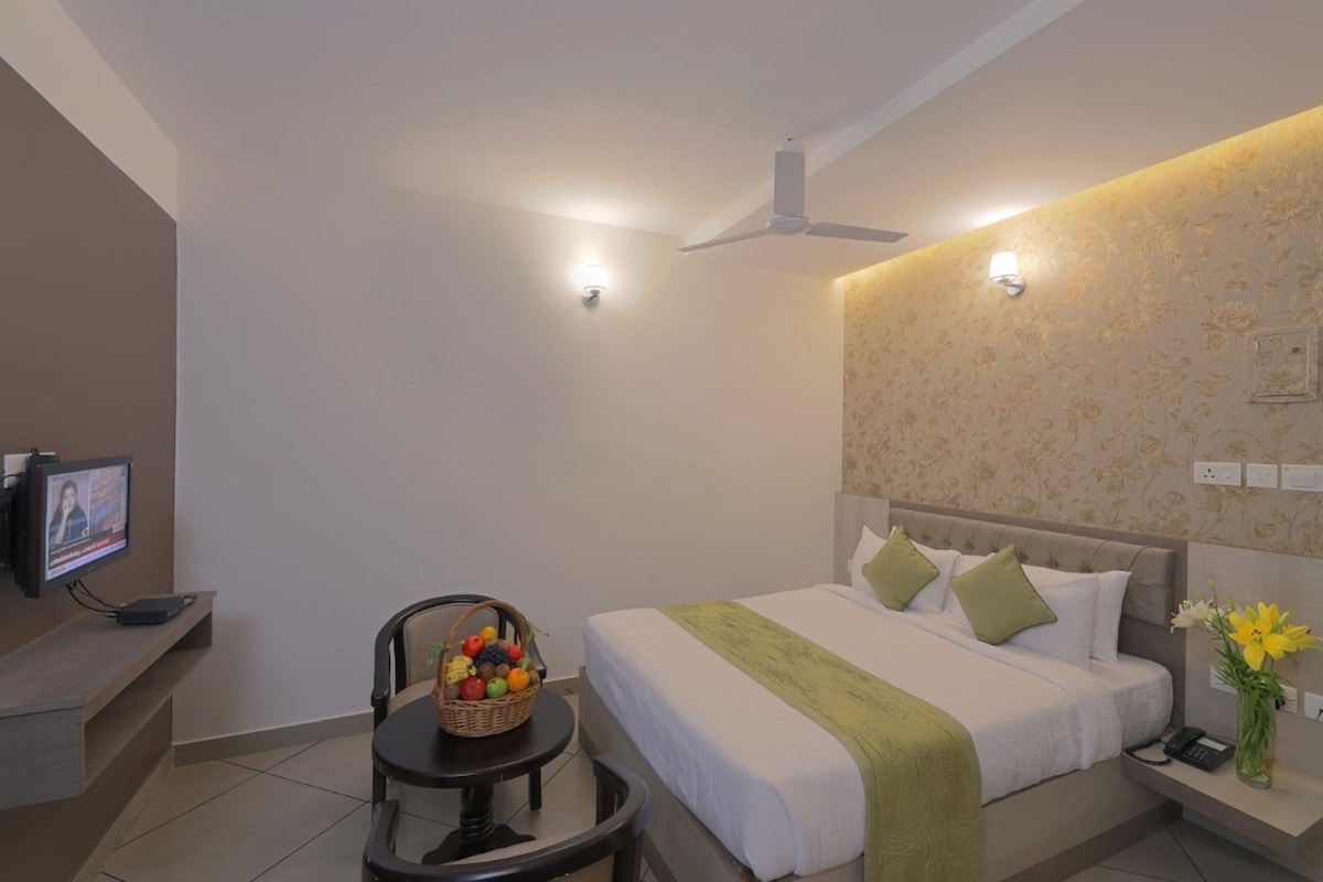 A hotel room with a bed, a desk with a television, a chair, a fruit basket, and a nightstand with a vase of flowers.