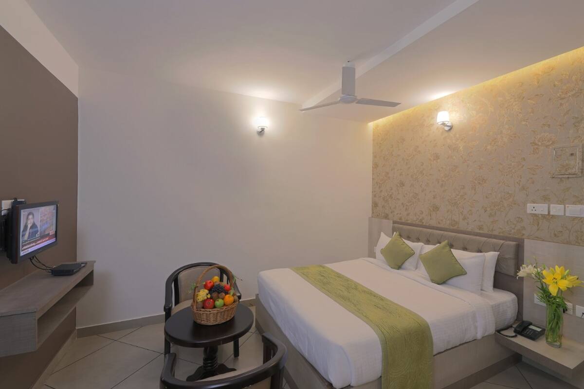 A hotel room with a bed, a desk with a computer, a chair, a fruit basket, and a vase with flowers.