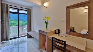 A hotel room with a balcony, a desk with a mirror, a vase with flowers, a coffee maker, and a water bottle. - Tea Castle Munnar (Devikolam)