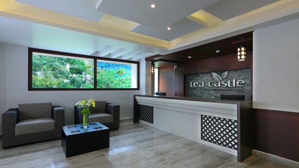 A reception area with a modern design, featuring a reception desk, two sofas, a coffee table with a vase, and a large window with a view of greenery. - Tea Castle Munnar (Devikolam)