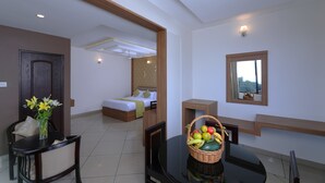 A hotel room with a bed, a dining table with chairs, a small kitchen area, and a window with a view. - Tea Castle Munnar (Devikolam)