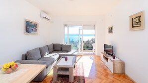 A modern living room with a grey sofa, a wooden coffee table, and a TV on a wooden stand. There is a large window with a view of the sea. - Apartment Marija (Novi Vinodolski)